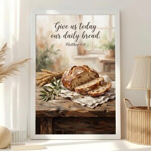 Puede incluir: Una impresión artística enmarcada que muestra una hogaza de pan crujiente con dos rebanadas sobre un paño blanco, acompañada de espigas de trigo y una rama de olivo. El texto dice "Give us today our daily bread. Matthew 6:11" en letra cursiva.