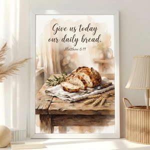 Può includere: Una stampa artistica incorniciata con un'illustrazione ad acquerello di una pagnotta di pane a lievitazione naturale affettata su un tavolo di legno. Il testo recita, Give us today our daily bread. Matthew 6:11. Un ramoscello d'ulivo e spighe.