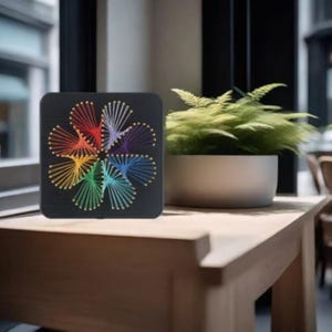 May include: A square black wooden board featuring a string art design of a multi-colored flower. The pattern is created with thin threads in shades of red, orange, yellow, green, blue, and purple, anchored by small metallic pins in a circular arrangement.