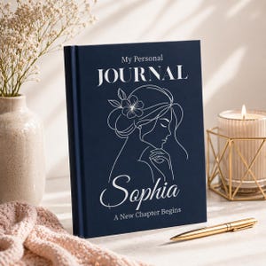 Puede incluir: Un diario azul marino con las palabras "My Personal JOURNAL" en la parte superior y el nombre "Sophia" en cursiva debajo de un dibujo lineal del perfil de una mujer. El texto "A New Chapter Begins" está en la parte inferior. Un bolígrafo dorado descansa sobre la superficie.