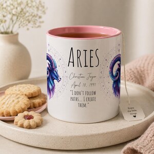 Puede incluir: Taza Aries de cerámica blanca con interior rosa, con una ilustración de carnero azul y morado. La taza incluye el nombre "Aries", una cita, un nombre y una fecha. Una bolsita de té con "Enjoy the little things" está adjunta.
