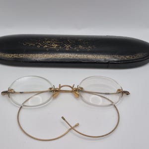 May include: Antique round eyeglasses with a gold-colored frame and clear lenses. The glasses are next to a black, oval-shaped case with gold detailing. The case is closed and appears to be made of leather.