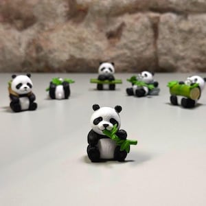 Könnte beinhalten: Eine Sammlung von Miniatur-Panda-Figuren in verschiedenen Posen. Die Pandas sind schwarz und weiß, einige halten grünen Bambus. Die Figuren stehen auf einer hellen Oberfläche, mit einer Backsteinmauer im Hintergrund.
