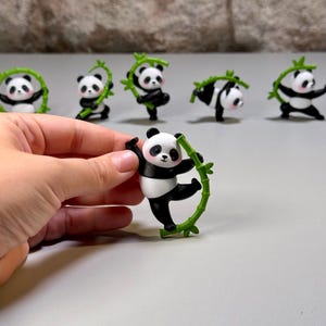Könnte beinhalten: Eine Sammlung kleiner Panda-Figuren, die jeweils einen schwarz-weißen Pandabären mit einem grünen Bambusstab zeigen. Die Pandas haben rosa Wangen und sind in verschiedenen Posen dargestellt, darunter stehend, kletternd und balancierend.