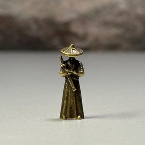 Könnte beinhalten: Eine kleine Messingfigur im Antik-Stil, die eine stehende Gestalt mit einem breitkrempigen konischen Hut und einem langen Gewand darstellt. Die Figur hält ein langes, schmales Schwert mit beiden Händen vertikal vor der Brust und hat ein metallisches Finish.
