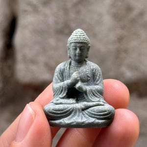 Könnte beinhalten: Eine kleine, graue, steinartige Buddha-Figur in sitzender Meditationshaltung. Die Figur weist eine texturierte Oberfläche, einen detaillierten Ushnisha auf dem Kopf und vor der Brust positionierte Hände auf. Die Skulptur ist kompakt.