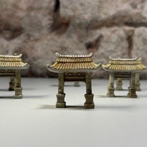 Könnte beinhalten: Miniatur-Nachbildungen von asiatisch anmutenden Strukturen mit gewölbten Dächern und detaillierten Säulen. Die Strukturen weisen ein verwittertes Aussehen mit Gold- und Grautönen auf. Der Hintergrund ist eine strukturierte, steinartige Oberfläche.