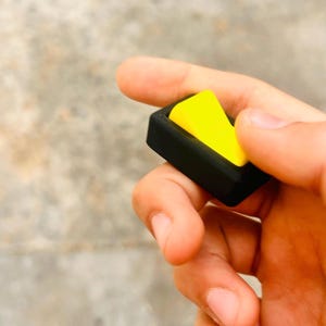May include: A black and yellow switch, held in a hand. The switch has a rectangular black frame with a bright yellow toggle. The image is a close-up, highlighting the contrast between the colors and the shape of the switch.