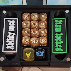 Könnte beinhalten: Ein schwarzer Spiel-Organizer aus Kunststoff mit Fächern für Würfel und Spielsteine. Der Organizer trägt die Texte "Ability Used" und "Item Locked". Er enthält braune Würfel mit weißen Zahlen und einen gelben Würfel mit Pikachu-Grafik.