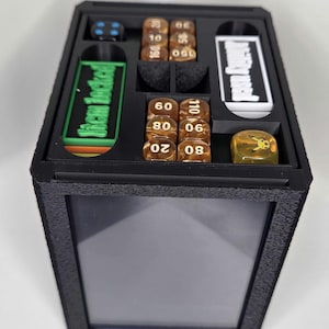 Könnte beinhalten: Eine schwarze, rechteckige Aufbewahrungsbox mit einer transparenten Front und einem Schaumstoffeinsatz für Spielzubehör. Enthalten sind braune nummerierte Würfel, ein blauer Würfel, ein gelber Würfel mit Pikachu-Motiv, ein grüner Anhänger mit Item locked.