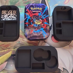 Könnte beinhalten: Schwarze, 3D-gedruckte Kartenhalter mit mehreren Fächern, darunter ein Fach für einen QR-Code. Eine Pokémon-Sammelkartenspiel-Box ist ebenfalls vorhanden. Die Box ist lila mit einer farbenfrohen Grafik eines Pokémon-Charakters.
