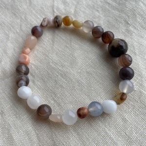 Pode incluir: Uma pulseira elástica composta por contas lisas, redondas e de formas irregulares em uma variedade de cores, incluindo branco, cinza translúcido, rosa suave, marrom escuro, bege e cinza carvão, dispostas em um padrão circular.
