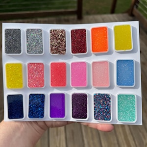 May include: A rectangular white sheet containing eighteen small, individual rectangular containers filled with various colored glitters. The colors include black, silver, bronze, red, orange, yellow, neon yellow, coral, pink, light pink, blue, dark blue, purple, violet, dark purple, multi-colored, and teal.