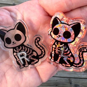 May include: Two cat-shaped resin charms featuring a black skeleton design. One charm is clear with a silver letter R, while the other is filled with iridescent orange glitter and features a silver letter L. Both display a cartoon cat silhouette.