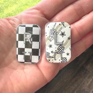 May include: Two rectangular, clear resin pieces. One features a black and white checkered pattern with a silver "R" and "TT". The other has a white background with stars, a skeleton hand, and a silver "L" and "TT".