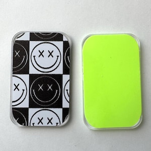 May include: Two rectangular phone grips. One features a black and white checkered pattern with smiley faces with X-shaped eyes. The other is a solid neon green color. Both have clear, rounded edges.