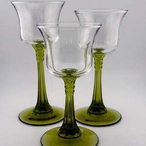 Peut inclure: Trois gobelets en verre transparent avec des pieds et des bases vert olive. Les bols des gobelets ont une forme évasée en forme de tulipe. Les tiges ont un design décoratif et texturé. Les bases sont rondes et plates. Les gobelets sont disposés sur un fond blanc.