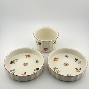 May include: A three-piece ceramic dish set. It features two round, shallow dishes and a small cup. Each piece is off-white with a scalloped edge and a floral pattern. The rims are a deep red.