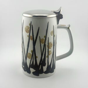 May include: A white ceramic beer stein featuring a textured black abstract pattern of vertical, pointed shapes and scattered gold-toned circular accents. The vessel includes a polished silver-colored metal lid with a thumb-operated hinge mechanism.