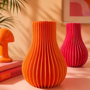 Op de afbeelding: Een feloranje vaas met een verticale, accordeonachtige geplooide textuur en een ronde basis die naar boven toe taps toeloopt. De vaas staat naast een kleinere oranje lamp en een tweede, identieke vaas in een levendige magentarode kleur.