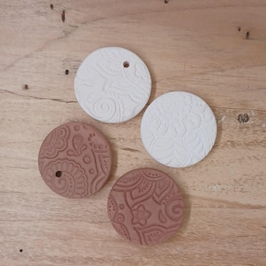 May include: Four circular clay pendants featuring intricate embossed patterns. Two pendants are white and two are terracotta-colored. Each piece has a small circular hole near the edge for hanging, with detailed floral and swirling designs.