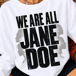 May include: A white crewneck sweatshirt featuring the black distressed text WE ARE ALL JANE DOE printed on the front. The design includes silhouettes of female profiles in a light grey colour positioned behind the bold black lettering.