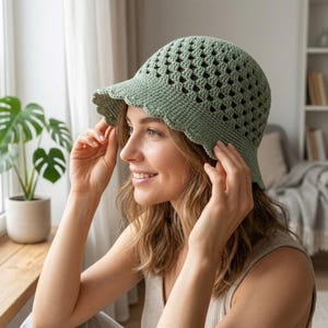 Op de afbeelding: Een saliegroene gehaakte bucket hat met een gestructureerd, opengewerkt ruitpatroon op de kroon en een stevige, geschulpte rand langs de boord. De hoed is gemaakt van garen met zichtbare steekdetails over het gehele ontwerp.
