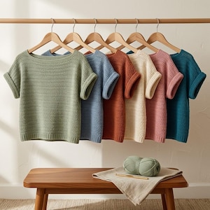 Peut inclure: Une collection de sept chemises en crochet à manches courtes suspendues sur des cintres en bois, alignées. Les chemises présentent un motif en maille texturée et sont disponibles en vert sauge, bleu, terre cuite, crème, vieux rose et bleu canard.