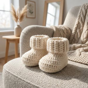 Peut inclure: Une paire de chaussons pour bébé crochetés à la main de couleur crème, dotés d'un revers côtelé épais. Les chaussons sont fabriqués à partir d'un fil épais avec un motif de points visible, offrant un aspect texturé et une forme arrondie.