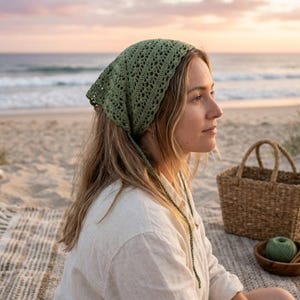 Peut inclure: Un foulard vert en crochet de forme triangulaire, noué à l'arrière. Le foulard présente un motif ajouré délicat. L'image montre le foulard sur une personne à la plage, avec un panier en osier et une couverture en arrière-plan.
