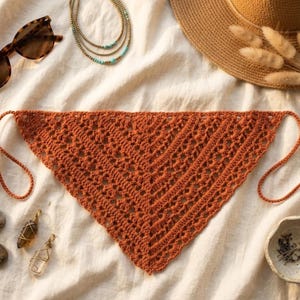 Peut inclure: Un foulard triangulaire au crochet de couleur rouille avec des attaches à nouer. Le foulard présente un motif ajouré et dentelle. Des lunettes de soleil, un chapeau de paille et des bijoux sont également visibles.