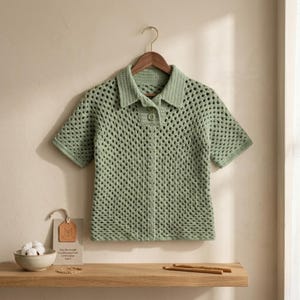 Peut inclure: Une chemise en crochet vert clair à manches courtes, avec un col et une fermeture boutonnée sur le devant. La chemise est suspendue à un cintre en bois. Le tissage ouvert du crochet crée un aspect texturé. La chemise est exposée sur une étagère en bois.