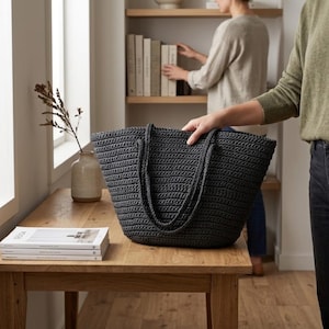 May include: A dark charcoal gray tote bag crafted with a thick, textured crochet knit pattern. The bag features two long, sturdy shoulder straps and a wide, open-top design, showcasing a consistent, dense stitch throughout its entire construction.