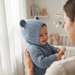 Peut inclure: Un pull pour bébé en crochet bleu poussiéreux doté d'une capuche avec deux petites oreilles d'ours arrondies sur le dessus. Le vêtement est réalisé avec un point texturé et comprend trois boutons bleus assortis sur le devant pour la fermeture.