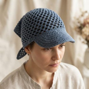 Peut inclure: Un couvre-chef en crochet bleu poussiéreux doté d'un motif en maille ajourée en forme de losange sur la calotte et d'une visière solide et finement tricotée. L'accessoire comprend deux liens triangulaires à l'arrière en fil bleu.