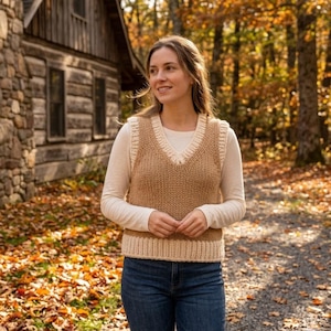 Peut inclure: Un gilet tricoté beige avec une bordure crème et un col en V. Le gilet est porté sur une chemise blanche à manches longues et associé à un jean bleu. L'arrière-plan présente une cabane et un feuillage d'automne.