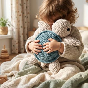 Peut inclure: Un enfant serre un jouet en peluche tortue crocheté à la main. La tortue a une carapace bleu clair, une tête et des membres beiges et des yeux noirs. Le jouet en peluche est doux et câlin.