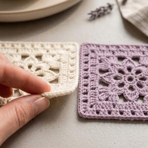 May include: Two square crochet coasters featuring a central floral motif with intricate openwork detailing. One coaster is off-white and the other is a soft lavender purple. The square pieces are crafted with fine yarn in a repeating geometric pattern.