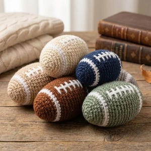 Peut inclure: Un ensemble de six petits jouets en crochet en forme de ballon de football, déclinés en beige, bleu marine, marron et vert sauge. Chaque objet en laine présente des lacets blancs cousus et des bandes blanches aux extrémités de l'ovale.