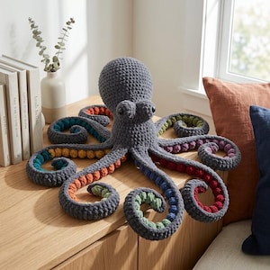 Peut inclure: Une grande peluche pieuvre crochetée grise avec huit tentacules enroulés. Chaque tentacule présente une doublure intérieure de couleur différente, notamment des tons orange, bleu sarcelle, jaune, vert, violet et rouge, avec une surface texturée.