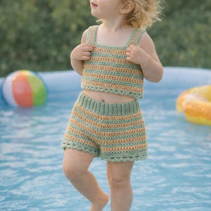 Peut inclure: Ensemble deux pièces au crochet pour enfant, composé d'un haut court et d'un short. L'ensemble est rayé vert clair et beige. L'enfant se tient dans une piscine bleue, avec un ballon de plage et une bouée gonflable.