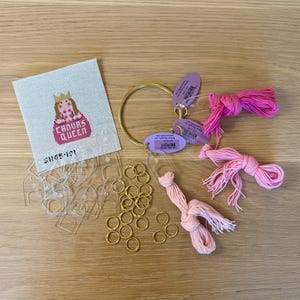 Op de afbeelding: Een vierkant borduurcanvas met een kruissteekontwerp van een figuur met een kroon en de tekst "CANVAS QUEEN" in het roze. Inclusief een gouden metalen ring, drie bundels roze borduurgaren, diverse doorzichtige plastic labels en kleine gouden ringetjes.