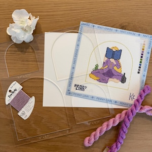 May include: A needlepoint kit with a cartoon image of a person reading a book, along with clear plastic templates, embroidery floss, and a white flower. The needlepoint canvas measures 4 x 4.25 inches.
