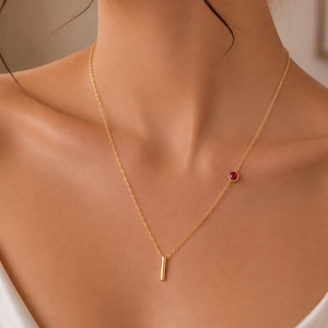 Op de afbeelding: Een gouden ketting met een kleine, verticale cilindervormige hanger in het midden en een enkele, ronde, rood ingezette edelsteen die asymmetrisch langs de ketting aan de rechterkant van de centrale staafhanger is geplaatst.