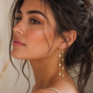 Peut inclure: Une paire de boucles d'oreilles pendantes dorées composées de deux chaînes fines suspendues à un petit clou. Une chaîne présente trois pierres transparentes en forme de poire, tandis que l'autre comporte trois petites pierres rondes.