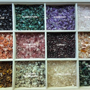 May include: A rectangular white tray with twelve compartments filled with small tumbled gemstones. The stones are labeled Black Tourmaline, Bloodstone, Amethyst, Rose Quartz, Sunstone, Strawberry Quartz, Garnet, Flower Agate, Tiger's Eye, Rainbow Moonstone, Clear Quartz, and Green Moss Agate. A small wooden scoop rests in the final compartment.