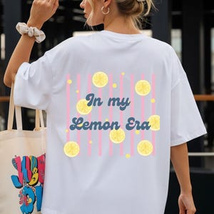 Könnte beinhalten: Ein weißes T-Shirt mit einem Rückendruck aus vertikalen rosa Streifen, gelben Zitronenscheiben und dem Text In my Lemon Era in dunkelblauer Schreibschrift. Das T-Shirt wird mit einer beigen Tragetasche mit buntem Grafikdruck kombiniert.