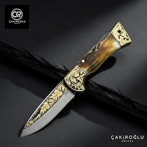 Puede incluir: Una navaja plegable con un diseño floral dorado en la hoja y el mango. El mango tiene un patrón marrón y crema. La navaja está etiquetada como "No 90". El logotipo de la marca "ÇAKIROĞLU KNIVES" es visible.