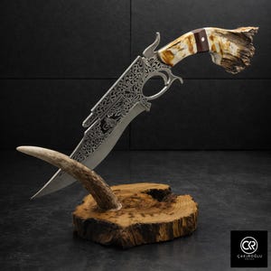 Puede incluir: Cuchillo plateado ornamentado con un mango decorativo de hueso y madera. La hoja presenta intrincados grabados y descansa sobre una base de madera con una cornamenta de ciervo. El cuchillo es un artículo decorativo.