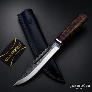 May include: A fixed-blade knife with a textured, dark metal finish on the upper blade and a polished edge. The handle is made of dark, patterned wood with two red bands. It is accompanied by a black leather sheath and a small black card with the text ÇAKIROĞLU BIÇAK MAĞAZASI.